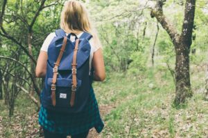 Blond woman with a backpack shown from behind hinking in the green woods.