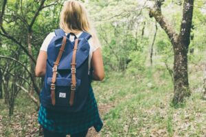 Blond woman with a backpack shown from behind hinking in the green woods.
