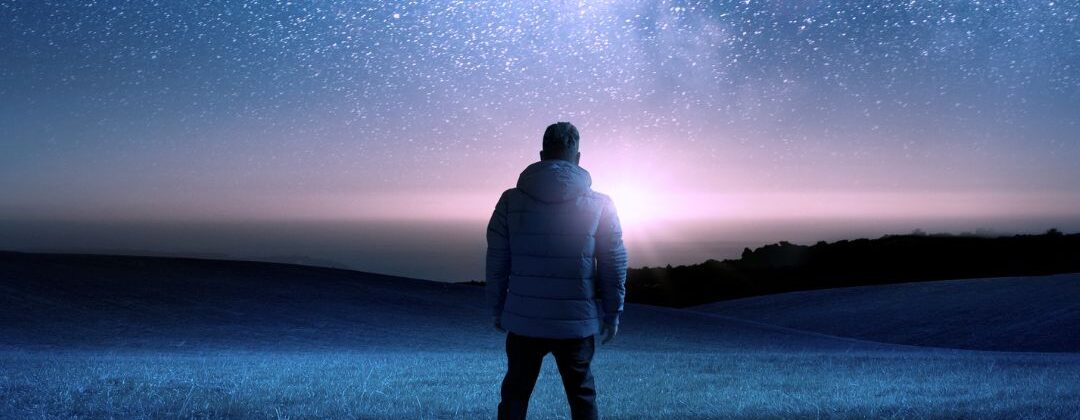 A person in a puffy white coat is seen from the back as they stare across a field at the galaxy of stars.